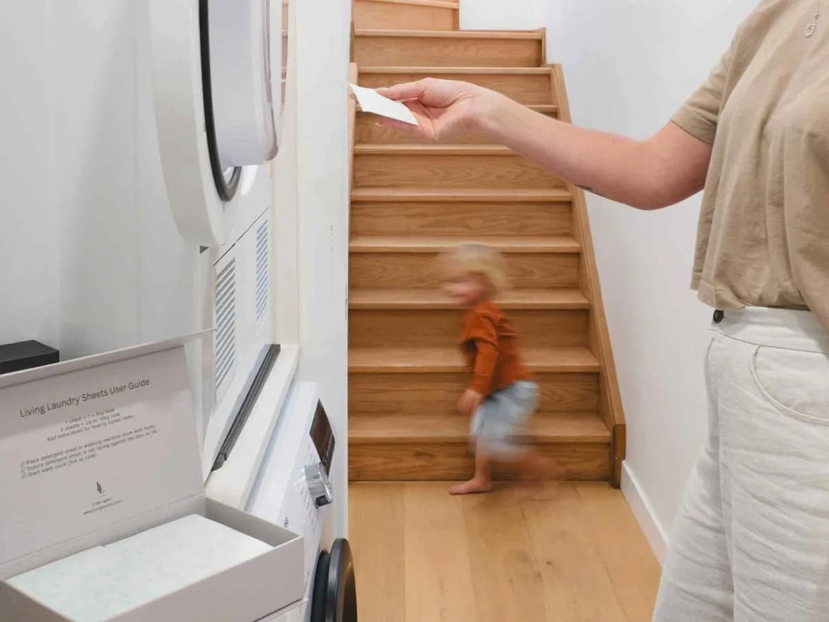 Person using a washing machine with detergent sheets with a child walking up a wooden staircase in the background.