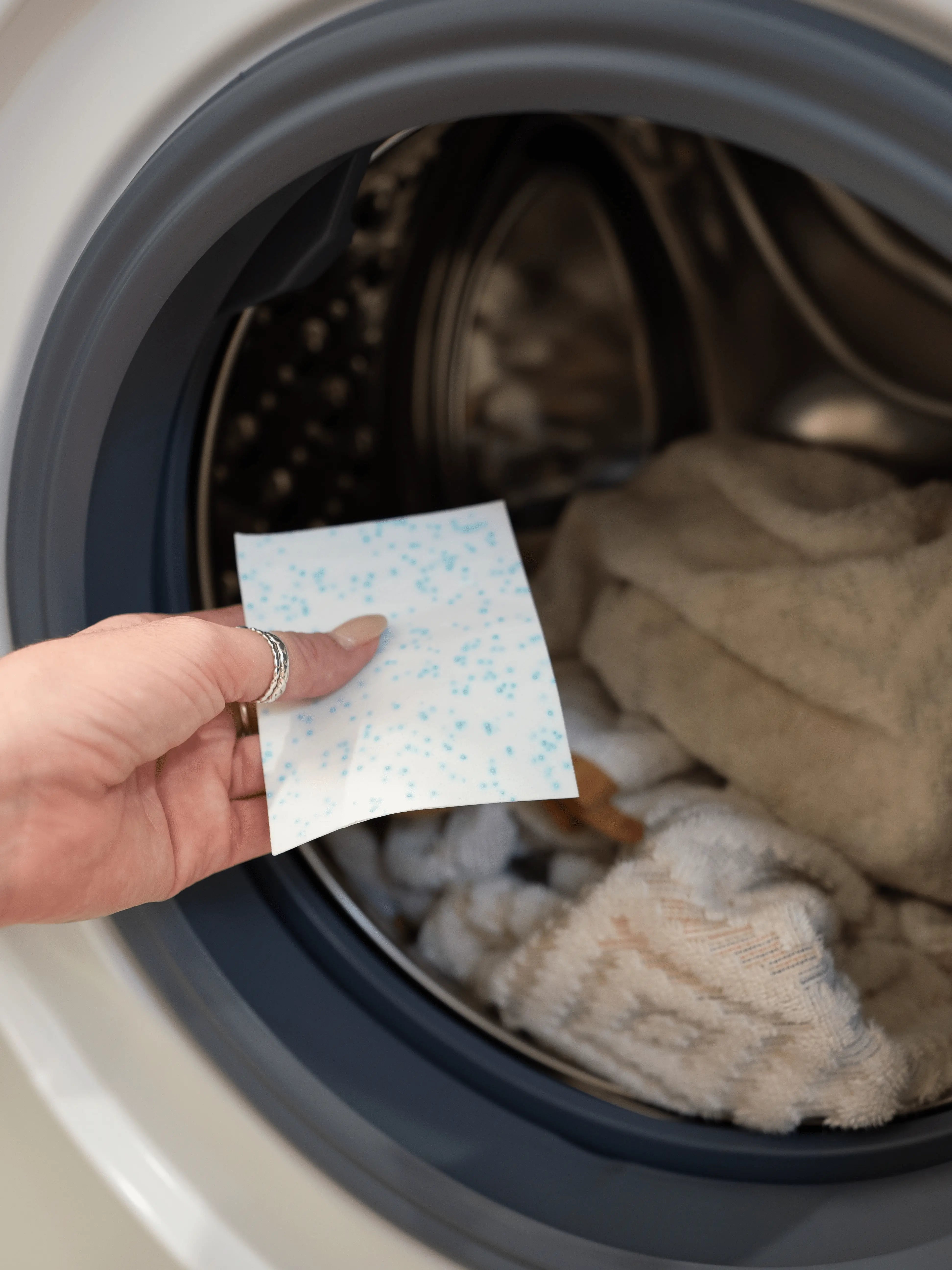 Hand holding a laundry detergent sheet in front of a washing machine with clothes inside.
